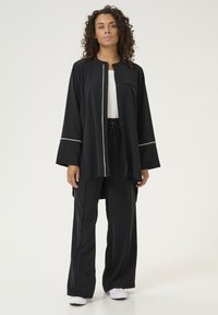 Melna, brīva piegriezuma jaka ar pogām priekšpusē un kontrastējošām baltām apmalēm, kombinēta ar atbilstošām melnām biksēm. Gluda, viegla auduma.