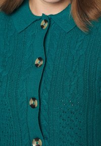 Teal stickad kavaj med kabelstickad design, krage och stora rundade knappar med kontrasterande mitt.