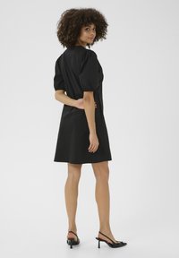 Crna haljina s kratkim napuhanim rukavima, uskim strukom i ravnom siluetom. Glatka tkanina s dužinom do koljena. Nosi se s crnim potpeticama.