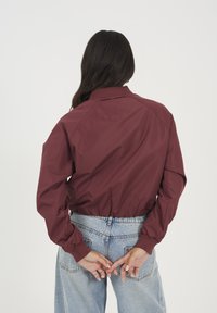 Veste bomber légère bordeaux avec un col et des poignets côtelés, mettant en avant un design court, une texture lisse et une taille ajustée.