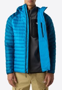 Blå puffjacka med quiltat mönster, huva, dragkedjeförsedd bröstficka och elastiska ärmslut, bärs över ett svart lager och tanfärgade byxor.