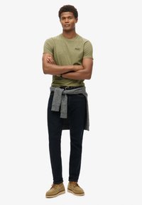T-shirt a maniche corte verde oliva, jeans scuri, maglione grigio a maglia legato in vita e scarpe beige con una finitura testurizzata.