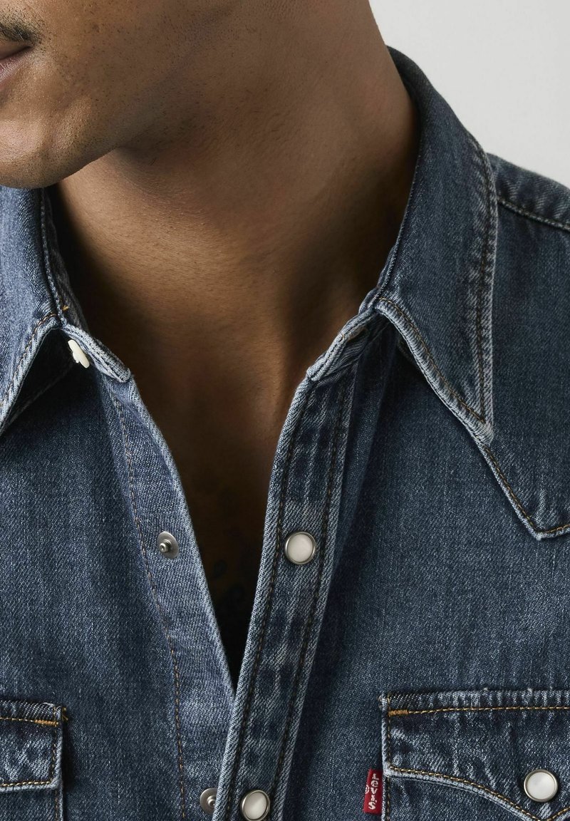 Mann trägt ein dunkelblaues Jeanshemd mit Knöpfen und Kragen, zeigt das untere Gesicht und den Hals vor einfachem Hintergrund.