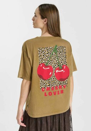 Femme tournée vers la gauche portant un t-shirt ample marron avec un imprimé léopard, deux grosses cerises rouges et un texte rouge "Cherry Lover" dans le dos.