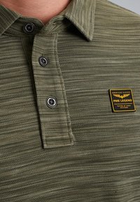 Polo vert olive en tissu texturé, présentant trois boutons noirs et un badge PME Legend jaune sur la poitrine gauche.