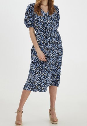 Kvinde iført en blå blomsterprint midi-kjole med pufærmer og bindebånd i taljen, kombineret med beige kilehælssandaler på en hvid baggrund.