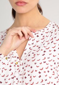 Vit blus med röd fjärilstrypt, mjukt tyg, knappdetaljer, V-hals och gathered accenter vid axlarna.