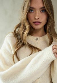 Kremkrāsas, pārāk liels adīts džemperis ar ribotām piedurknēm, apaļu kakla izgriezumu un vienu pogu; mīksta, teksturēta dzija.