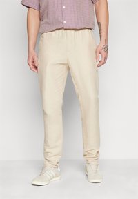 Lichtbeige op maat gemaakte broek van zachte stof met een elastische tailleband. Draagt een paars geruit overhemd met korte mouwen en grijze sneakers.