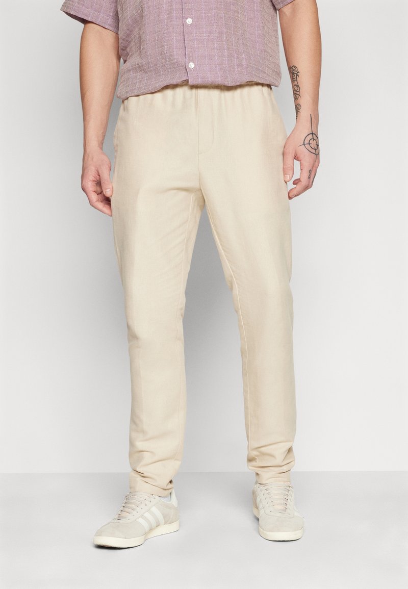 Lichtbeige op maat gemaakte broek van zachte stof met een elastische tailleband. Draagt een paars geruit overhemd met korte mouwen en grijze sneakers.