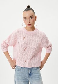 Różowy sweter robiony na drutach z wzorami w warkocz, z luźnymi rękawami i kilkoma małymi przetarciami. Noszony z jasnoniebieskimi dżinsami.