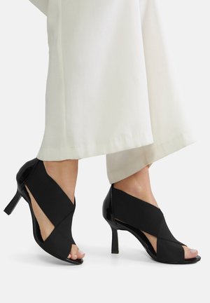 Fekete magassarkú cipők, nyitott orral, rugalmas anyagból készült, keresztezett dizájnnal és fényes lakk díszítéssel a sarkon.