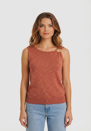 Jeune femme aux cheveux ondulés portant un haut sans manches en maille couleur rouille avec des boutons en bois, et un jean bleu clair, debout devant un fond blanc.