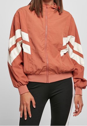 Personne portant une veste zippée couleur rouille avec des bandes en chevrons blanches sur les manches et un pantalon noir avec une bande latérale blanche.