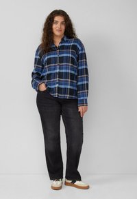 Woman stands against gray wall wearing blue plaid shirt, black jeans, and cream sneakers with black and brown details.