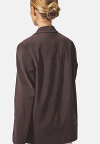 Brun kavaj med strukturerade axlar och en enda slits baktill. Har en smidig tygtyp och knäppta ärmslut.