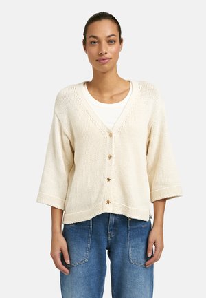 Woman wearing a cream knitted cardigan with three-quarter sleeves and decorative buttons over a white top and blue jeans.