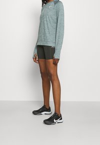 Langärmliges, teal-farbenes Sportshirt mit strukturiertem Finish, kombiniert mit dunklen grünen Shorts und schwarzen Nike-Sneakern mit weißen Akzenten.
