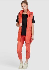 Orangen gesteppte Weste mit hohem Kragen, mit Reißverschlusstaschen; kombiniert mit einem schwarzen T-Shirt und dazu passenden orangen Slim Pants, weißen Sneakers.