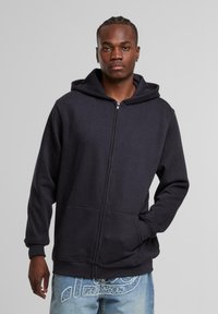 Sudadera con cremallera negra, capucha forrada, bolsillos frontales y puños acanalados. Textura de tela suave, con un diseño simple y minimalista.