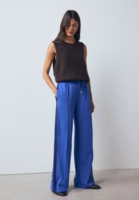 Barna, ujjatlan, kötött pulóver laza, kék szabadidőnadrággal, oldalcsíkkal és húzózsinórral. Fekete cipő egészíti ki a megjelenést.