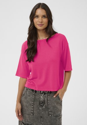 Žena s dlhými tmavými vlasmi, oblečená v jasnoružovom krátkym rukávom tričku a čiernej sukni s vysokým pásom v štýle acid wash, s veľkými prednými vreckami.