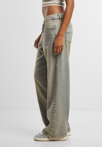 Person wearing loose, faded light blue wide-leg jeans with sneakers and a striped cropped top, standing sideways on a plain white background.