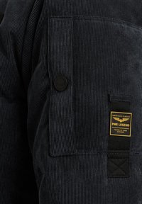 Chaqueta de pana azul oscuro con un bolsillo prominente que tiene un botón negro y una etiqueta de marca amarilla. La textura es acanalada y texturada.