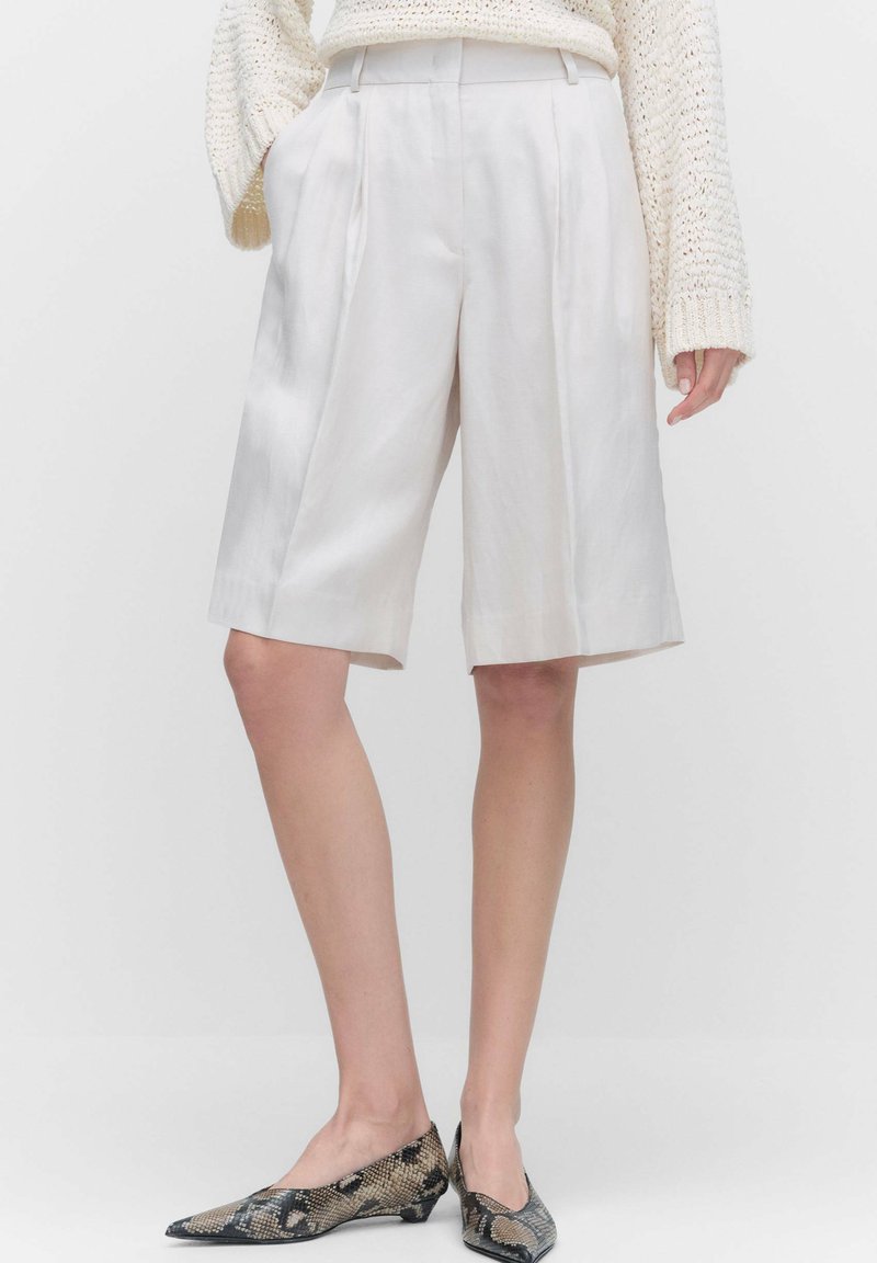 Ljust beige, högmidjade shorts i ett mjukt tyg med veck. Har en rak passform och fåll som når kn�äet, i kombination med mönstrade skor.
