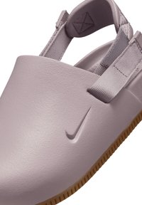 Lilla vormitud suss tekstuurse pinnaga ja kontrastse tan värvi kummitald; omaduseks reguleeritavad hallid rihmad ja Nike logo.