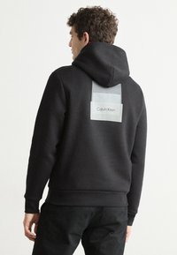 Czarny kaptur z miękkiego materiału, z jasnoszarym prostokątnym naszywką z logo "Calvin Klein" na plecach.