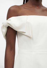 Άτομο με σκουρόχρωμο δέρμα που φοράει λευκό φόρεμα με ακάλυπτους ώμους, μεγάλο φιόγκο στο αριστερό μανίκι και εκτεθειμένες κλείδες.