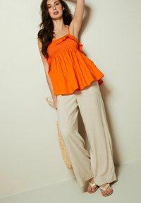Heldere oranje katoenen peplum tanktop met gegolfde randen, gecombineerd met lichtbeige linnen wijde broek en roomkleurige sandalen. Geweven strooien tas tentoongesteld.