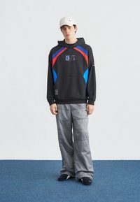 Jeune personne portant un sweat à capuche noir avec des accents bleus et rouges, un pantalon gris ample, des chaussures noires et une casquette blanche, debout sur un tapis bleu.