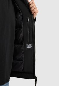 Zwarte jasinterieur met gewatteerde voering, ritsvak en een logo-patch. Gladde stof met een matte afwerking en zachte textuur.