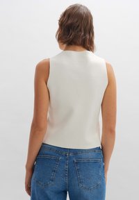 Kvinna med vågigt brunt hår bär en ärmlös vit topp och blå högmidjade jeans, fotograferad bakifrån mot en enkel ljus bakgrund.