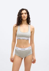 Γυναίκα με μακριά σγουρά μαλλιά που φοράει γκρι αθλητικό σουτιέν Calvin Klein και αντίστοιχα σλιπ, στέκεται μπροστά σε απλό λευκό φόντο.