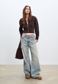 Felpa corta con zip di colore marrone sopra una maglietta bianca, abbinata a jeans a gamba larga di colore blu chiaro. Una borsa marrone scuro con una texture è appoggiata su una spalla.