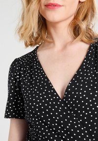 Czarna sukienka kopertowa w białe kropki, z krótkimi bufkami i głębokim dekoltem w kształcie litery V. Miękka, gładka tkanina z kwiatowymi detalami tekstury.