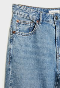 Jeans de mezclilla azul claro con una textura ligeramente desgastada, diseño de cinco bolsillos, remaches de cobre y detalles de costura visibles.