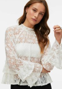 Blusa blanca de encaje con cuello alto, mangas largas acampanadas y bajo con volantes superpuestos. El encaje presenta patrones florales y una textura semitransparente.