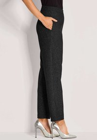 Vrouw in zwarte, gestructureerde pantalons met rechte pijpen, hand in de zak en zilveren, puntige hakken, staande zijwaarts.