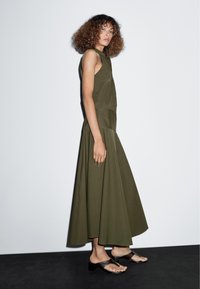 Olijfgroen mouwloos jurkje met knoopsluiting aan de voorkant en een uitlopende rok, gecombineerd met zwarte instappers, tegen een lichte achtergrond.
