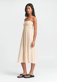Beige midi-kjole med smocket buste, tynde spaghetti-stropper og en svajende nederdel. Parret med sorte flip-flops. Glat stoftekstur.