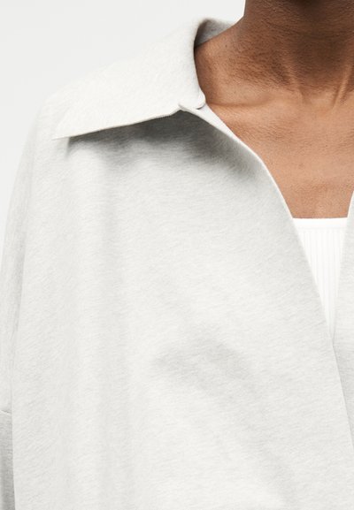 Κοντινό πλάνο ατόμου που φοράει ανοιχτό γκρι παλτό με γιακά πάνω από λευκό ριγωτό τοπ, εστιάζοντας στον λαιμό και το άνω μέρος του στήθους.