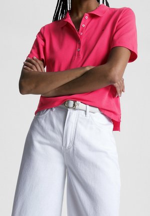 Person mit verschränkten Armen, trägt ein leuchtend pinkes kurzärmeliges Poloshirt, das in hoch taillierte weiße Hosen mit weißem Gürtel gesteckt ist.