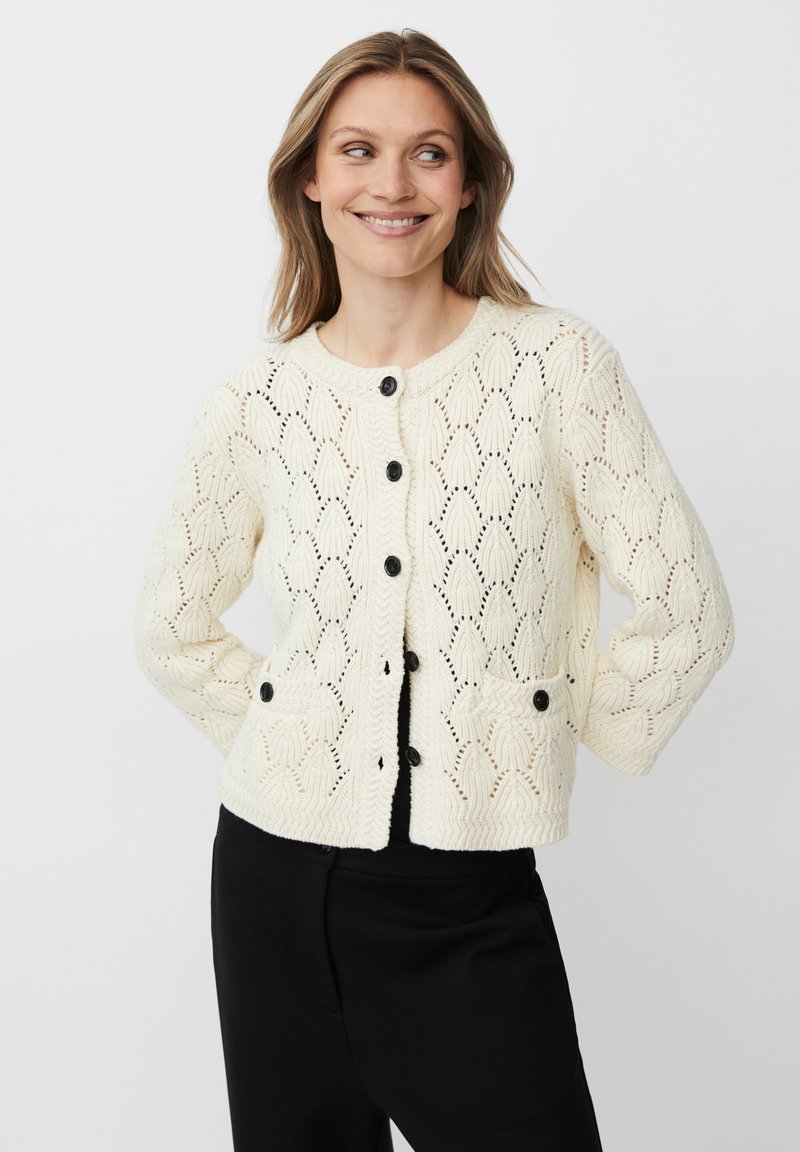 Donna che sorride e guarda di lato, indossando un cardigan di maglieria color crema con bottoni neri e pantaloni neri, su uno sfondo bianco semplice.