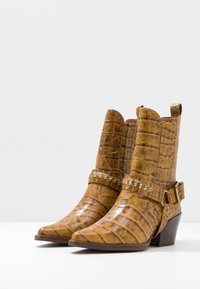 Botas de tobillo de cuero con relieve de cocodrilo en color mostaza, con un tacón grueso y una correa decorativa con acentos metálicos.