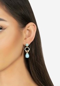 Pendientes en tono dorado con un diseño circular negro, adornados con un colgante de gota azul claro y acentos de piedras preciosas transparentes.