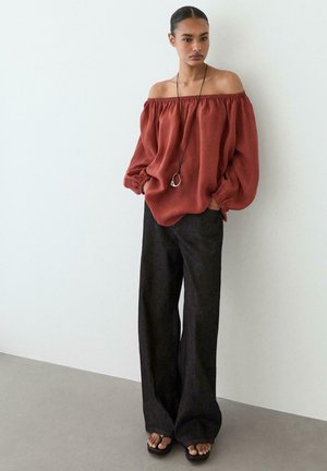 Kvinde iført en løs, rustfarvet off-shoulder top, vidde mørke bukser, sorte sandaler og en lang halskæde, står med hænderne i lommerne.